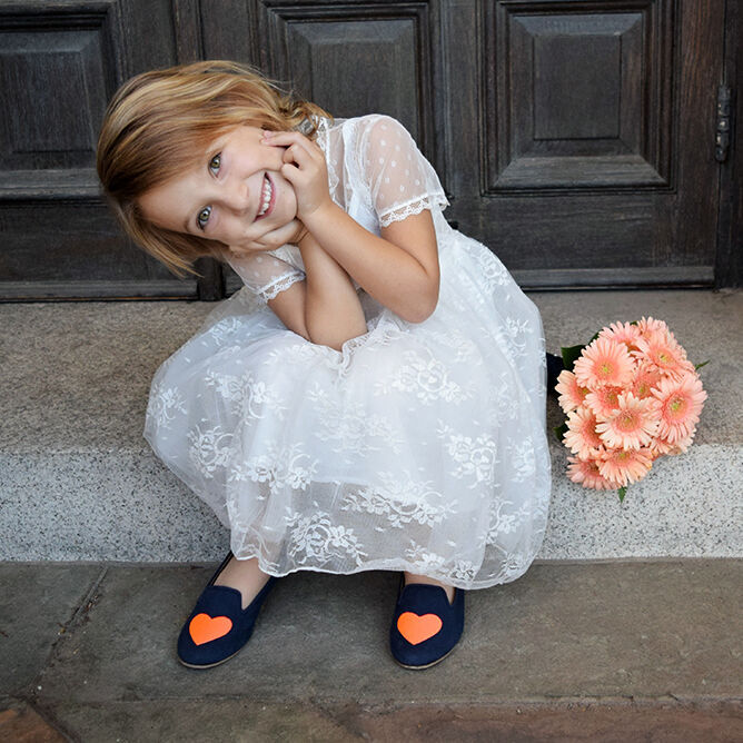 Ein Mädchen in einem hübschen weißen Kleid steht mit gefalteten Händen und einem Lächeln auf der Treppe. Sie trägt Schuhe mit Herzchen und sitzt neben einer Blume.