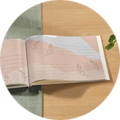 Ein aufgeschlagenes CEWE FOTOBUCH mit sanften, hellen Seiten und zarten Blumenmustern. Es liegt auf einem hellen Holztisch.
