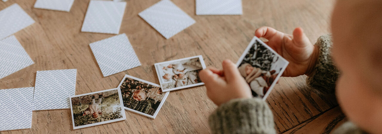 Ein Kind hält Fotos in der Hand und schaut darauf. Um das Kind herum liegen weitere Fotos auf einem Holzfußboden.