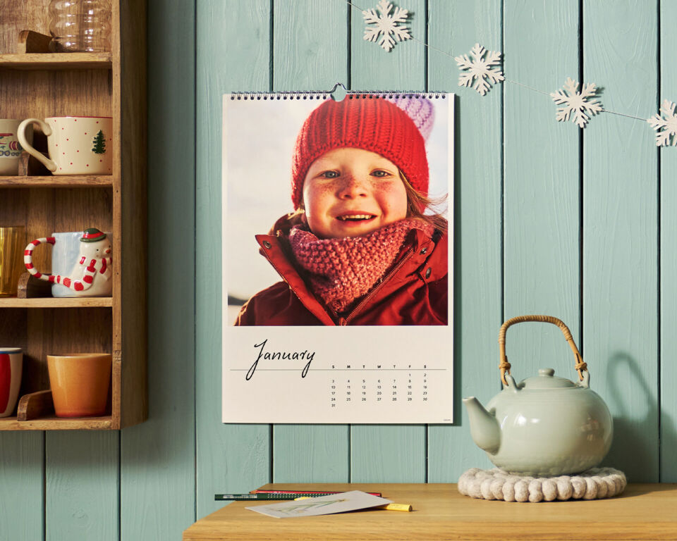 CEWE Wandkalender mit winterlichem Kinderfoto an türkiser Holzwand, daneben ein Tassenregal und eine Teekanne.