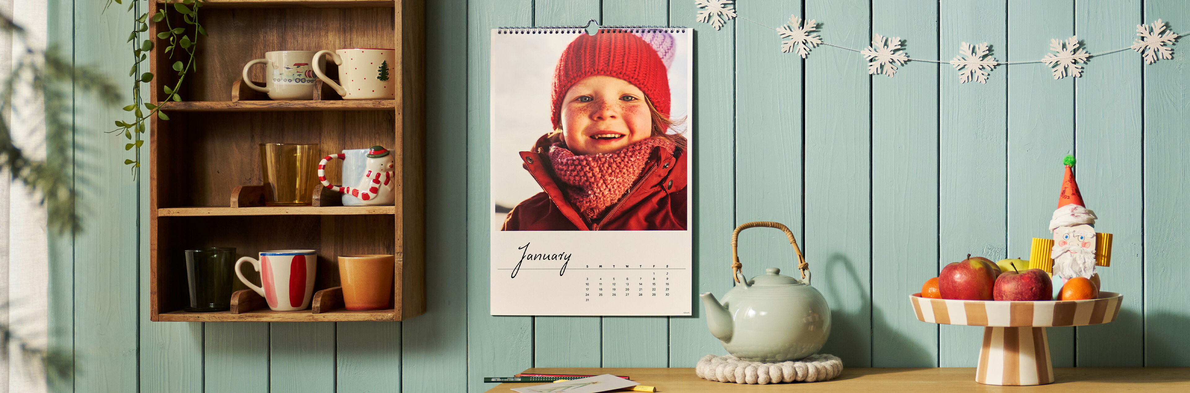 CEWE Wandkalender mit winterlichem Kinderfoto an türkiser Holzwand, daneben ein Tassenregal und eine Teekanne.