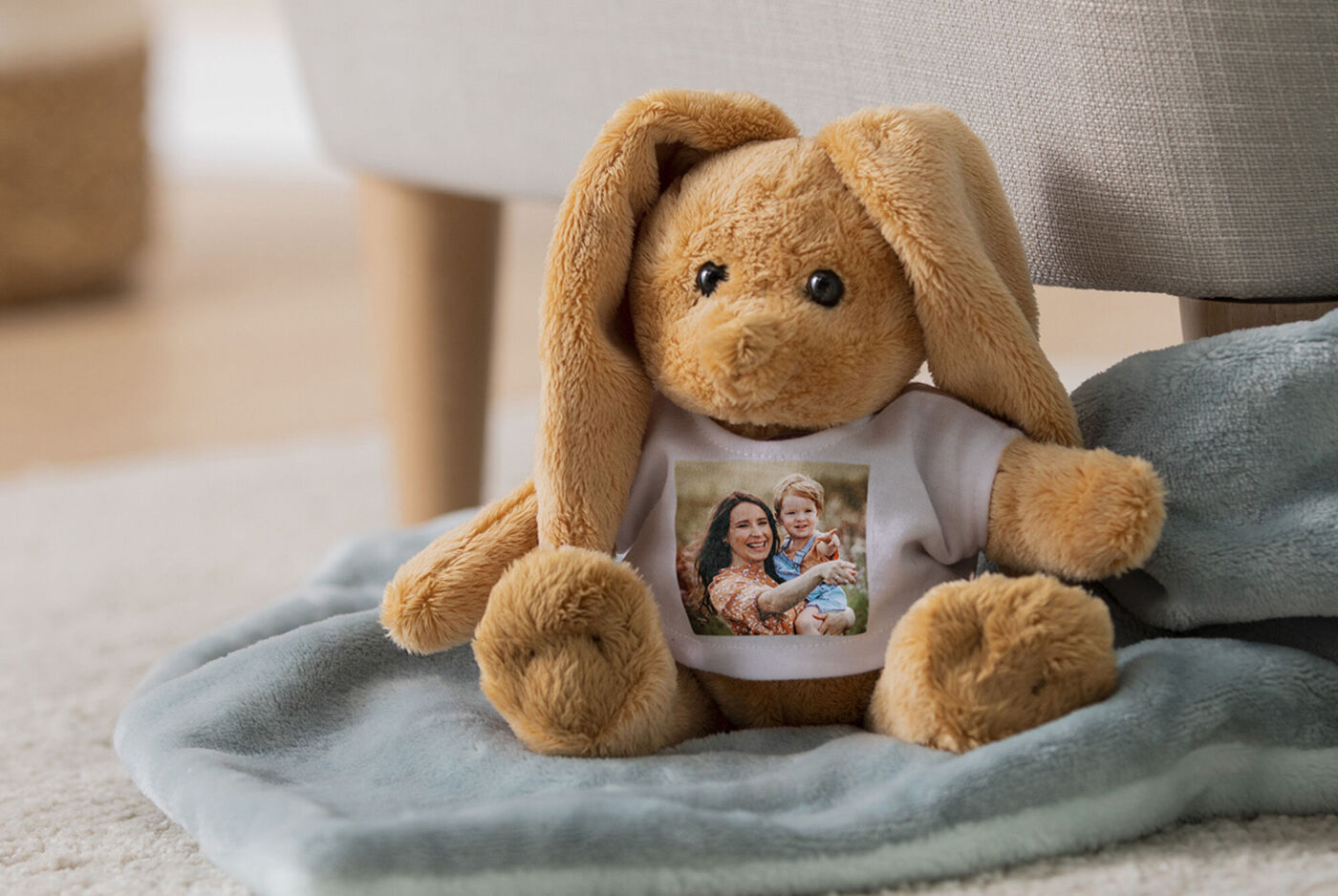 Ein niedlicher, plüschiger Hase mit einem T-Shirt, auf dem ein Foto von zwei Personen abgebildet ist, sitzt auf einer weichen Decke.