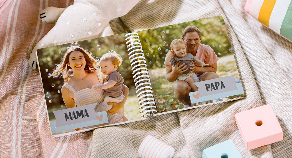 Das Bild zeigt ein offenes CEWE FOTOBUCH mit einer liebevollen Familienfotoszene. Auf der linken Seite ist eine Mutter mit ihrem Kind und auf der rechten Seite ein Vater, der ebenfalls sein Kind hält. Die Umgebung ist grün und sonnig.