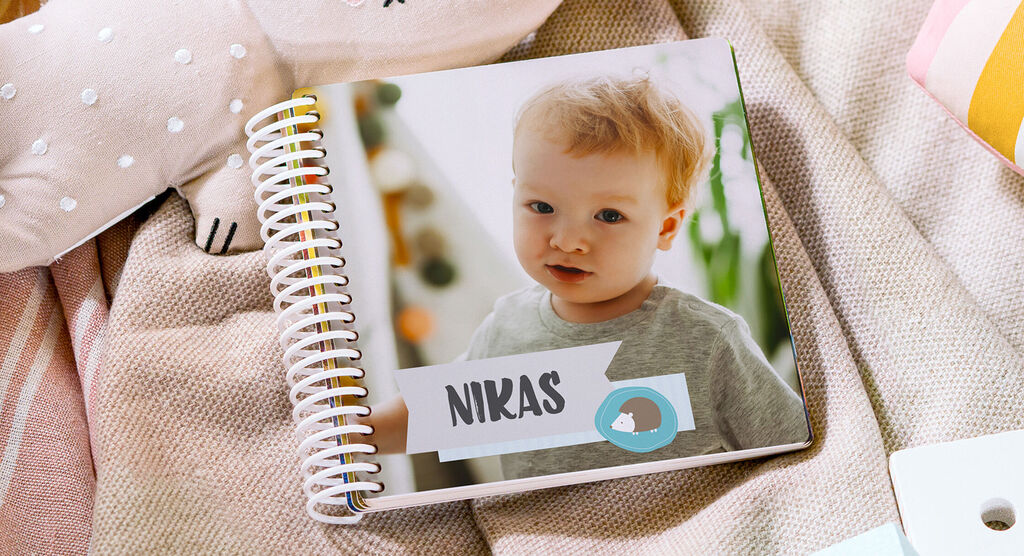 Ein CEWE FOTOBUCH auf einer Decke, umgeben von Spielsachen. Es hat den Namen 'Niklas' auf dem Cover.