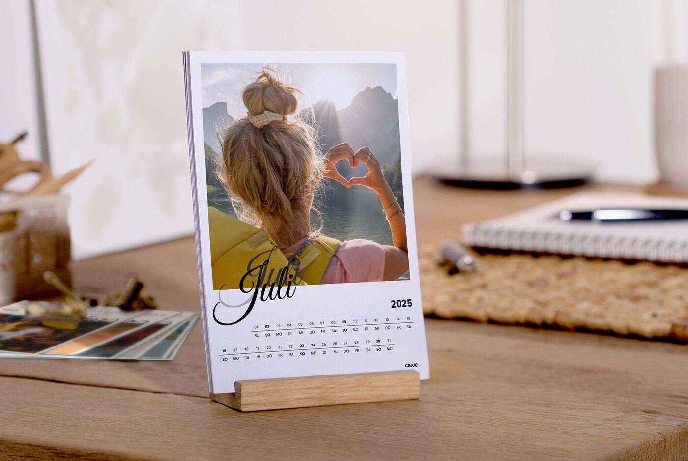 Eine Person mit lange Haare macht ein Herz mit den Händen vor einer malerischen Landschaft, ein personalisierter Kalender steht auf dem Tisch