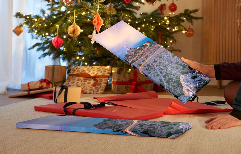 Eine Person hält ein großes Wandbild von einer Küstenlandschaft vor einem geschmückten Weihnachtsbaum und Geschenken.