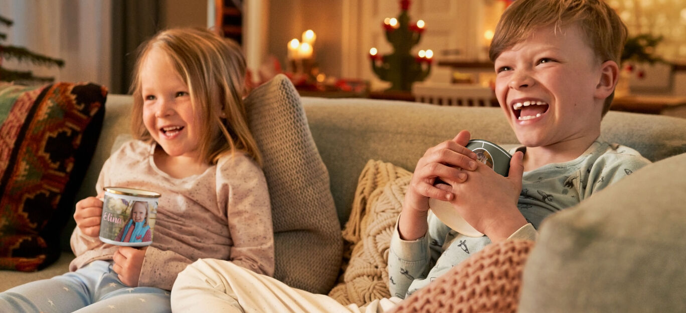Zwei Kinder entspannen auf einer Couch und halten Tassen in den Händen. Die Atmosphäre strahlt Gemütlichkeit aus.