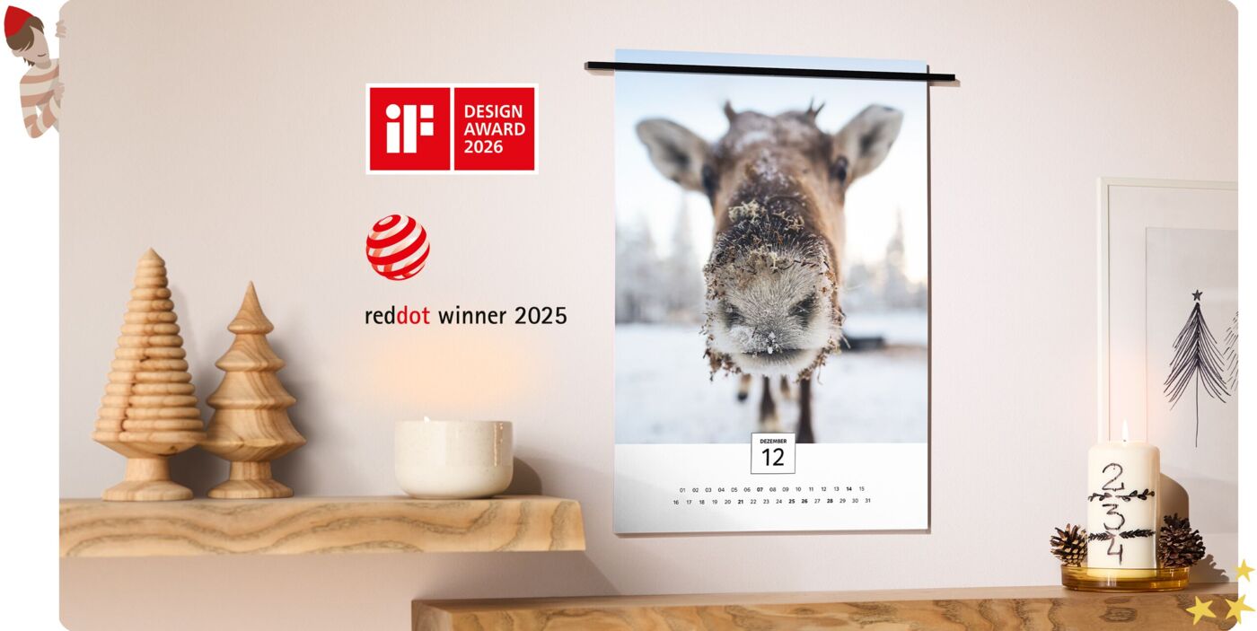 CEWE Fineline Wandkalender mit Rentierfoto im Winter hängt an Wand, umgeben von Weihnachtsdeko, iF Design Award 2026 und Red Dot Winner 2025