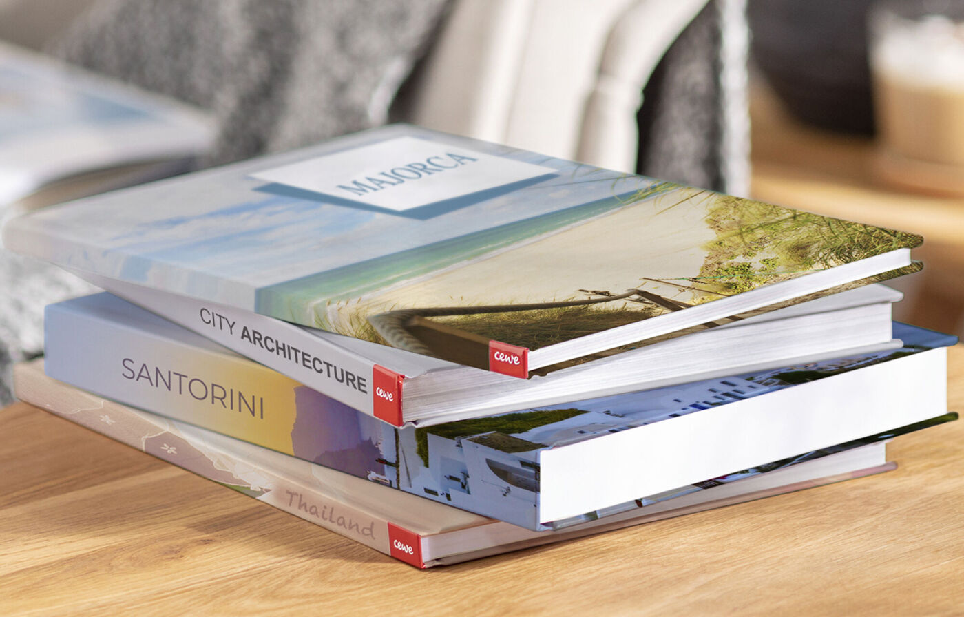Three beautifully designed photo books on a table. One shows a beach titled 'MAROCA'. Another presents city architecture with the title 'CITY ARCHITECTURE'. The third book is labeled 'SANTORINI'.