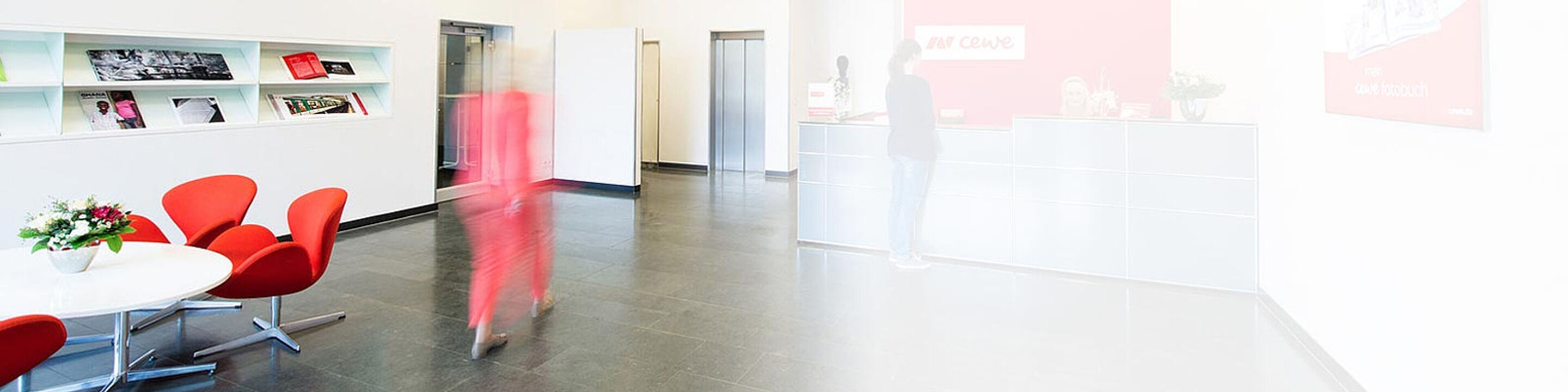 A person in a red dress walking in a modern reception area with red chairs and a reception desk.